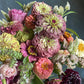 A vibrant, multicolored bouquet of freshly harvested flowers, including pink, yellow, white, and burgundy zinnias with green foliage, held by a hand against a gray concrete floor.